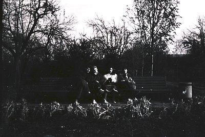 Silhouetted group of three seated around a small campfire in a wooded area, silhouetted against low light. Mid-20th century o...