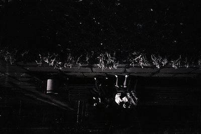 High-angle black-and-white shot of a snow-covered rooftop with intricate metal piping and gutters. Snow accumulates unevenly,...
