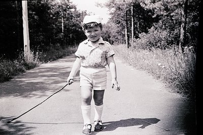 Mid-20th century boy in vintage attire—short-sleeved polo shirt, knee-length shorts, and knee-high socks—walks a leash with a...