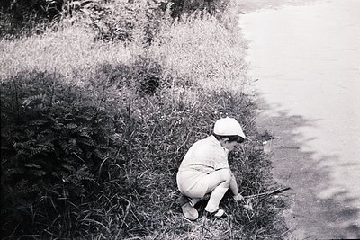 Mid-20th century black-and-white photo of a child crouching by a waterfront, holding a small object. Lightweight summer attir...