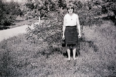 Vintage black-and-white portrait of a woman in a grassy outdoor setting, likely 1950s–1960s. She wears a short-sleeved blouse...