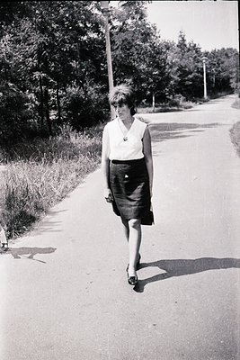 Mid-century woman in a sleek black-and-white dress walks along a tree-lined road, holding a small handbag. Short hair, V-neck...