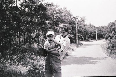 Two children pose on a rural roadside, likely mid-20th century (1950s–1960s). The boy in overalls carries a bag, while the yo...