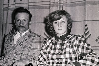Black-and-white portrait of a man and woman in matching plaid shirts, seated indoors. The man wears a mustache and a checkere...