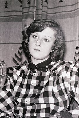 Portrait of a woman in a 1970s-style checkered blouse with a black collar, set against striped wallpaper. Her short, layered ...