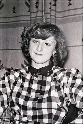Portrait of a woman in a 1960s-style checkered blouse, short bob haircut, and subtle makeup. Indoor setting with patterned wa...