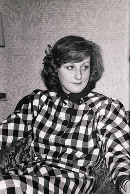Portrait of a woman in a plaid shirt, seated indoors with a patterned wallpaper background. Her short, voluminous hairstyle a...