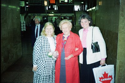 Three women pose indoors near an airport baggage claim, 1980s. Center figure wears a red blazer with a blue scarf, flanked by...
