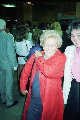 Vintage candid shot of an elderly woman in a bright red raincoat, smiling warmly while holding a blue garment. Indoor setting...