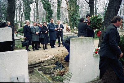 A solemn funeral procession in a cemetery, likely late 20th century. Group of mourners in formal attire—dark suits, scarves, ...