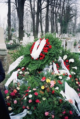 Elaborate floral tribute on a grave, featuring a large wreath with red roses, white lilies, and greenery, adorned with a whit...