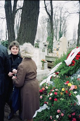 Two women in 1970s-era coats stand at a graveside adorned with floral tributes and a wreath. The gravestone features classica...