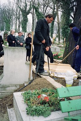 A man in formal attire tends to a grave in a cemetery, surrounded by mourners. The grave features a wreath and a small wooden...