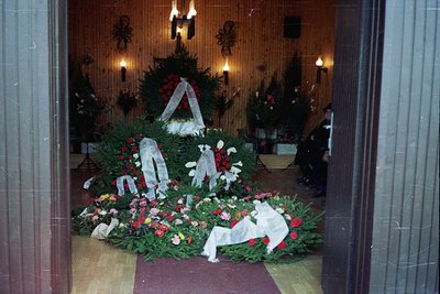 Elaborate funeral wreaths draped in greenery, white ribbons, and red roses surround a central memorial table in a dimly lit i...