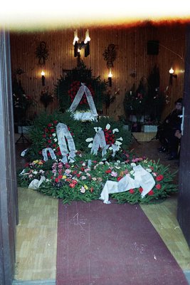 Memorial wreaths and floral tributes arranged in a solemn indoor display, likely a funeral or memorial hall. Wood-paneled wal...