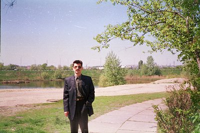 Man in dark blazer poses by a serene lakeside path, surrounded by greenery and paved walkway. Soft focus suggests late 1990s–...