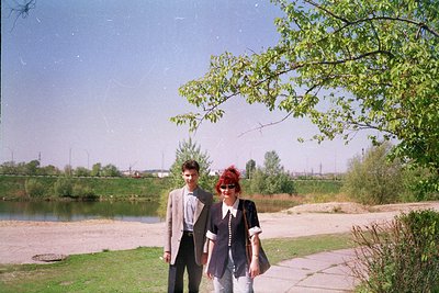 Vintage photo of two individuals posing outdoors beside a serene lake, framed by lush greenery and a tree. The man wears a li...