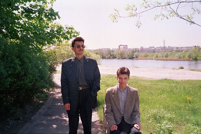 Two men pose by a riverbank in a park setting, likely late 20th century. The man on left wears a dark blazer, striped shirt, ...