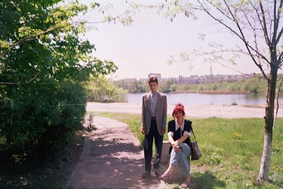 Two individuals pose on a paved riverside path, framed by greenery and a distant urban skyline. Man in suit, woman in skirt w...