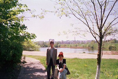 Two individuals in 1970s-style attire pose by a riverbank, surrounded by lush greenery and blooming trees. The man wears a su...
