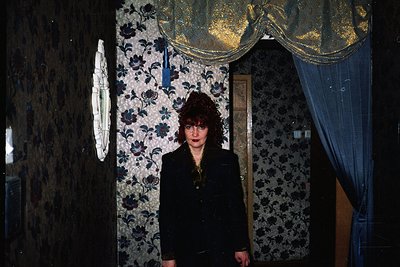 Vintage interior portrait featuring a woman in a dark blazer, standing in a dimly lit hallway with floral wallpaper and a gil...