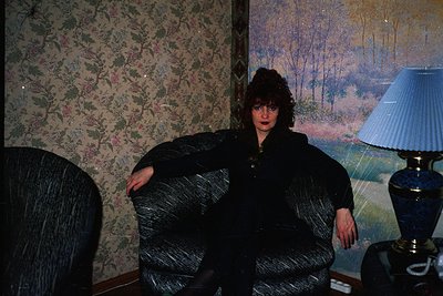 Vintage interior featuring floral wallpaper and a woman in a voluminous, draped black coat. Side table with ornate brass lamp...