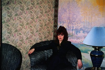 Vintage interior portrait featuring a woman in a dark, textured blazer leaning on a striped armchair. Floral wallpaper with m...