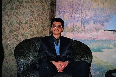 Vintage portrait of a man in a formal black suit with a blue tie, seated on a tufted armchair against a floral-patterned wall...
