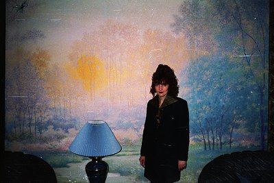 Portrait of a person standing in front of a large, abstract landscape mural with muted tones of blue, green, and orange. The ...