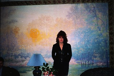 Portrait of a woman in a dark blazer standing before a large, abstract landscape mural depicting a sunset over trees. Blue ta...