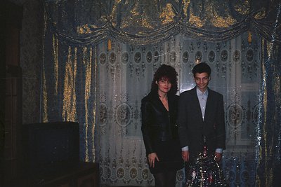 Two individuals pose in a dimly lit, ornate interior with intricate wallpaper featuring floral and geometric patterns. The ma...