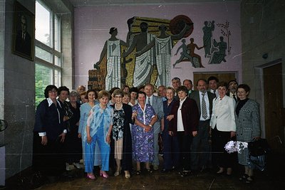 Group portrait in a Soviet-style interior, likely 1970s–1980s Eastern Bloc. Indoor setting features mural depicting industria...
