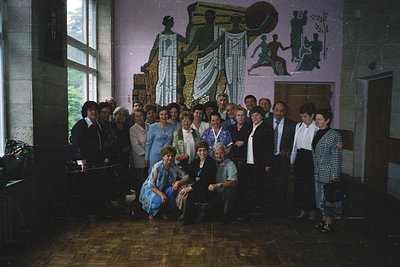 Group portrait in a grand hall featuring socialist-era mural depicting industrial workers and agricultural themes. Formal att...