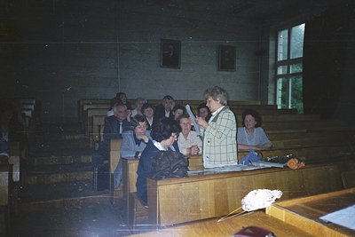 Vintage indoor gathering in a modest lecture hall or classroom, featuring tiered wooden benches. A woman in a checkered blous...