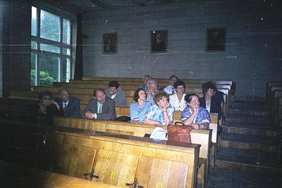 Vintage indoor lecture hall with tiered wooden benches, featuring a diverse group of seated adults in formal attire (1960s-70...