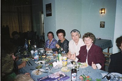 Family gathering in a modestly decorated indoor setting, likely a 1980s–1990s Eastern European home. Six adults seated around...