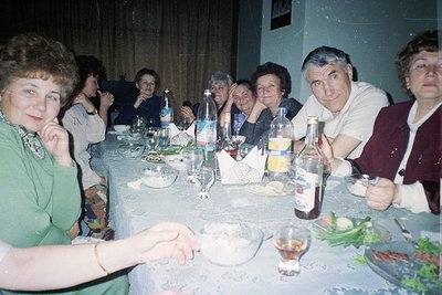 Vintage group dinner in a dimly lit indoor setting, likely Eastern Europe, 1970s–1980s. Round table with bottles of mineral w...