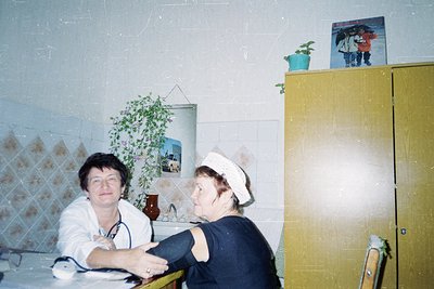 Vintage clinical exam room featuring a nurse in white uniform and stethoscope examining a patient in a blue sweater. Decor in...