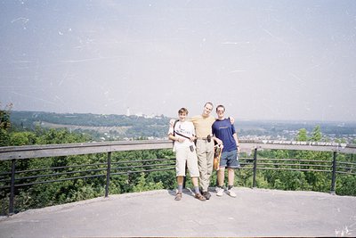 Three individuals pose on a high viewpoint platform with metal railings, overlooking a forested valley and distant cityscape ...