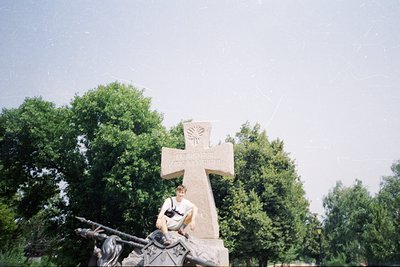 Monumental stone cross with Soviet-era hammer-and-sickle emblem at top, set in a park-like environment. A person poses beside...