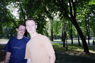 Two young men pose in a lush, shaded park setting, likely 1990s–2000s. The man on the left wears a dark blue graphic tee with...