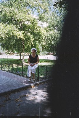 Woman in a white hat and sleeveless black top stands on a paved pathway beside a metal railing, holding a white garment. Lush...