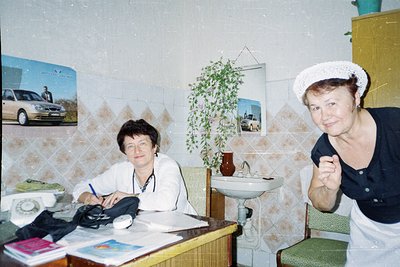 Two women in a modest indoor setting, likely a 1990s-era home. The woman on the left writes at a wooden desk with a rotary ph...