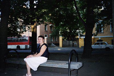 Vintage urban portrait of a woman seated on a metal bench under leafy shade, dressed in a sleeveless black top and white skir...