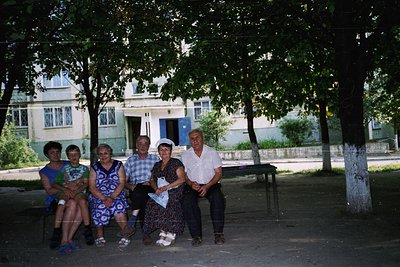 Group of five posing outdoors in a tree-lined courtyard, likely Eastern Europe, 1970s–1980s. Light-colored building with blue...