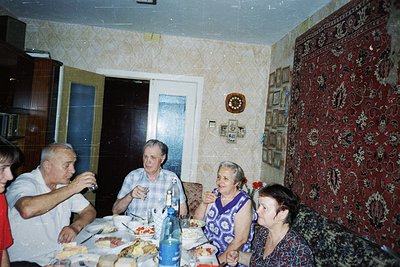 Four adults seated around a table in a 1970s-era living room, enjoying a meal. Patterned wallpaper with floral motifs and a r...
