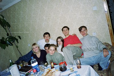 Vintage group portrait in a dimly lit indoor setting, likely a 1990s home gathering. Six individuals pose on a couch and tabl...