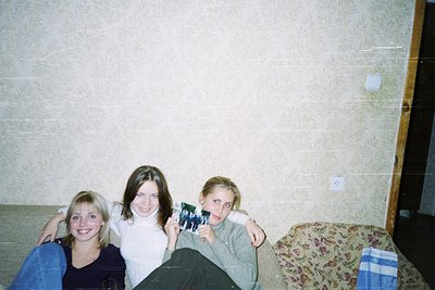 Vintage indoor portrait of three young women posing on a patterned sofa in a plain room. Center figure holds a black-and-whit...