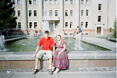 Two individuals pose on a stone ledge beside a tiered fountain in a Soviet-era urban plaza, likely late 20th century. The man...