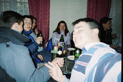 Indoor gathering of six adults in casual 1990s attire, seated around a table with beer bottles and glasses. Warm lighting and...